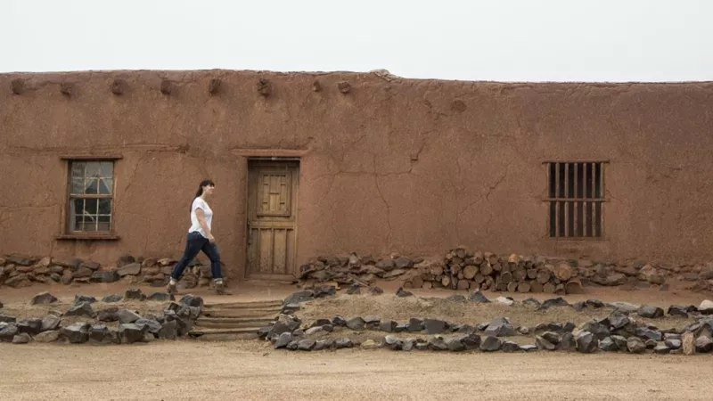 Woman walking on a path of stones in front of an old adobe building in Taos Pueblo.