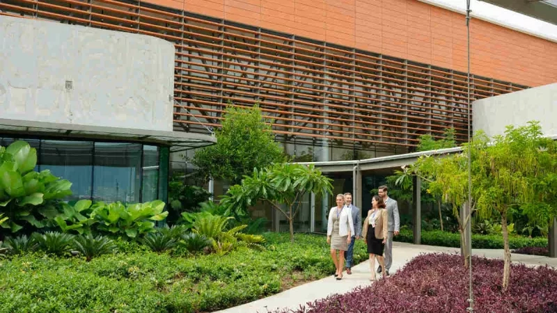 People walk on a path in front of a building with landscaping and an orange and gray facade.