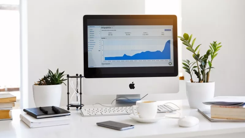 A white desk with an Apple monitor, keyboard, mouse, coffee, and potted plants on it.