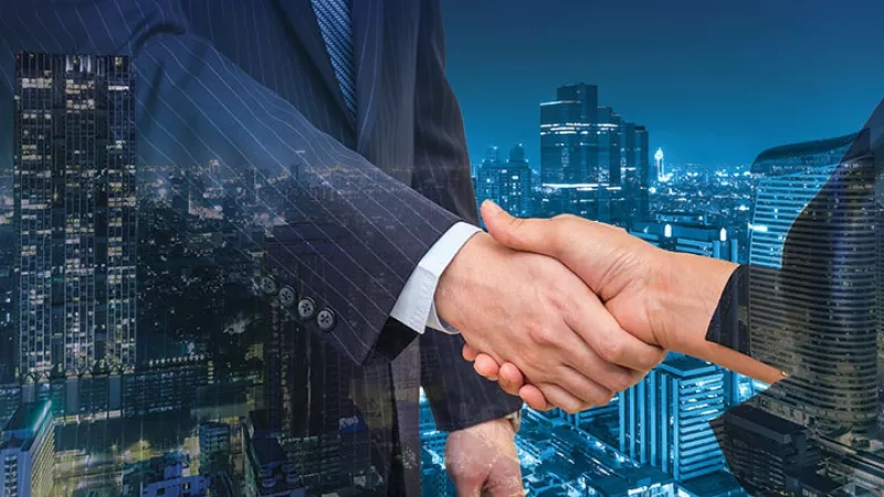 Two businessmen shake hands against a backdrop of a city skyline at night, symbolizing a business deal.