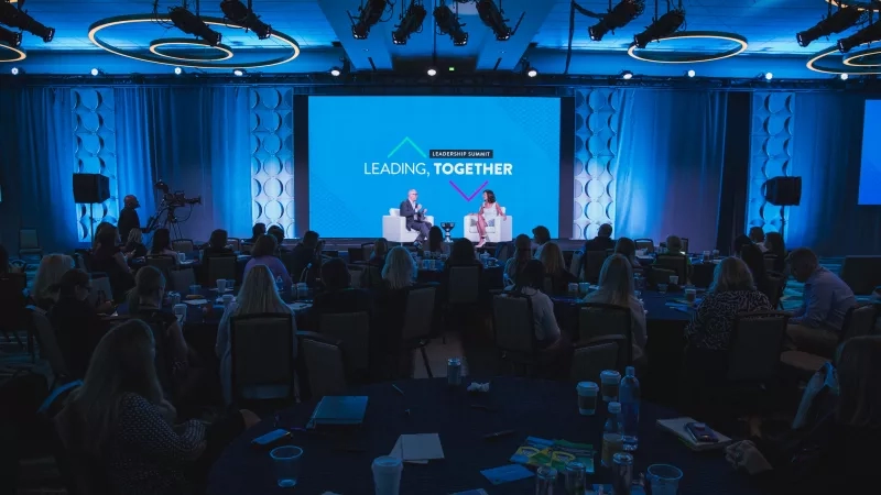 Leadership Summit is underway with a large group of people sitting at tables in front of a large screen.