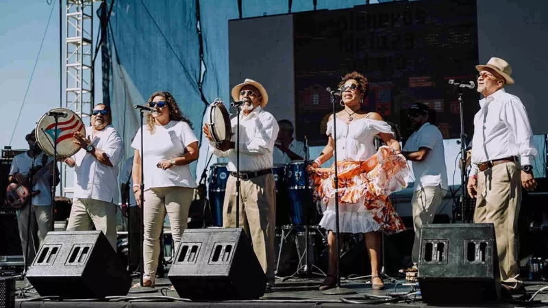 A group of musicians are performing on a stage with instruments and microphones in an outdoor concert.