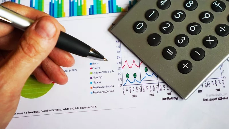A hand holds a pen next to a calculator on top of a financial document.