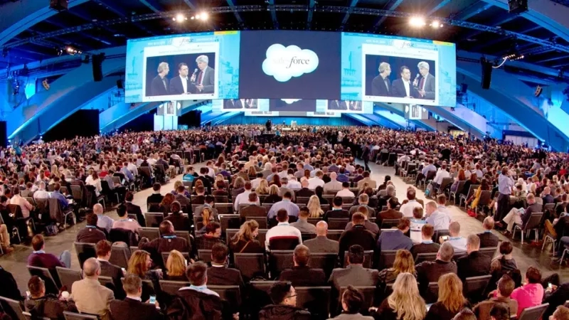 Salesforce conference with large screens showing speakers and a large audience seated in rows of chairs.