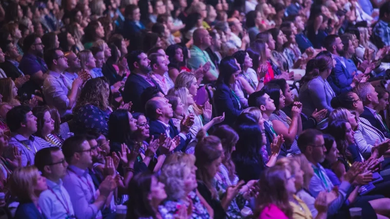 Crowd of people at a conference, many wearing green name tags, clapping in an auditorium.