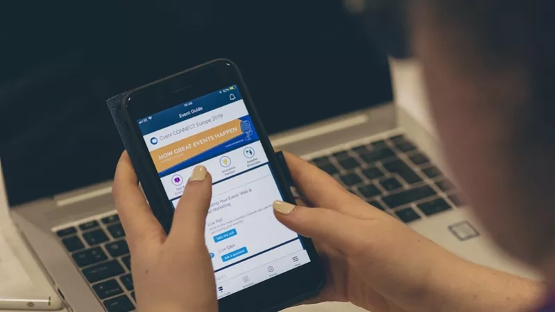 Woman holding a smartphone with the Event Connect Europe app open in front of a laptop on a table.