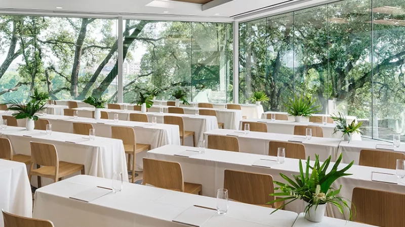 Many tables and chairs are arranged for a meeting in the room, with glass windows and trees outside.