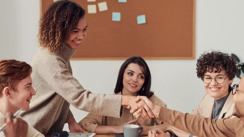 Group of people sitting indoors and a woman greeting another person with a handshake.