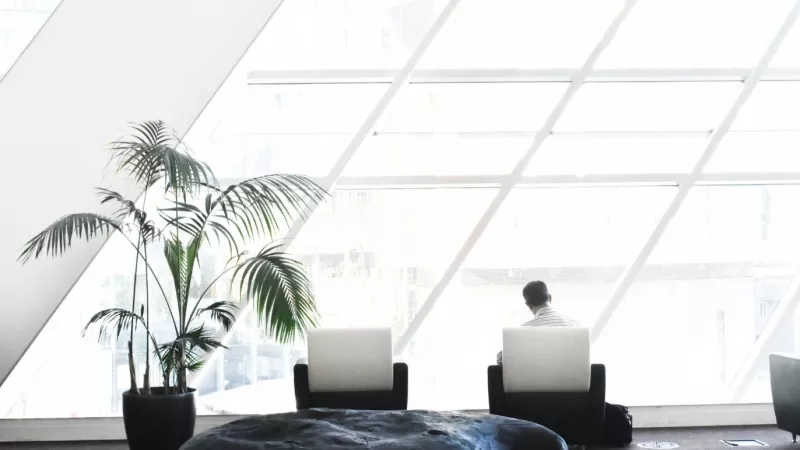 A man sitting on the chair inside the lobby with a potted plant next to him.