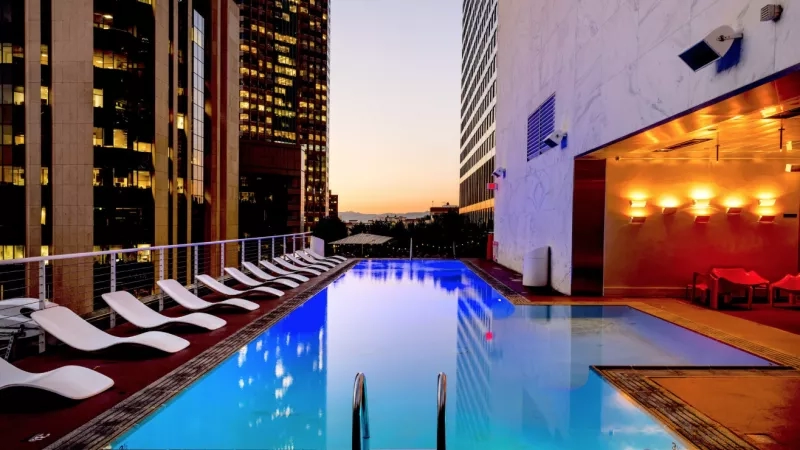 The swimming pool is surrounded by lounge chairs and tall buildings in the background at dusk.