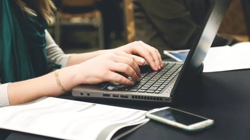 Woman types on laptop with cell phone and notebook next to her on table in a conference room.