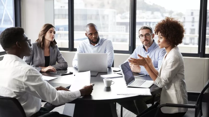 Four people in a meeting room with a glass window, discussing and gesturing while laptops are on the table.