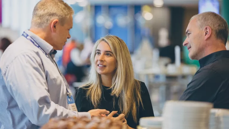 Three people are talking in a cafeteria with plates and bread on the table in the background.