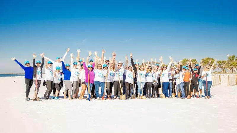 A group of people are standing on the beach holding shovels and wearing blue hats and shirts.