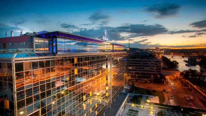 Aerial view of the glass-fronted Hilton Prague with the city and river in the background at dusk.