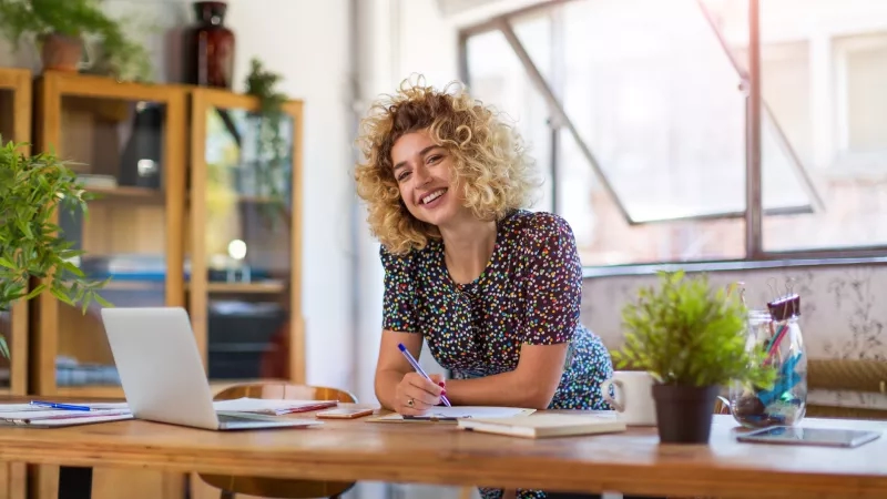 A smiling woman sitting at a desk with a laptop, pen, paper, and potted plants.