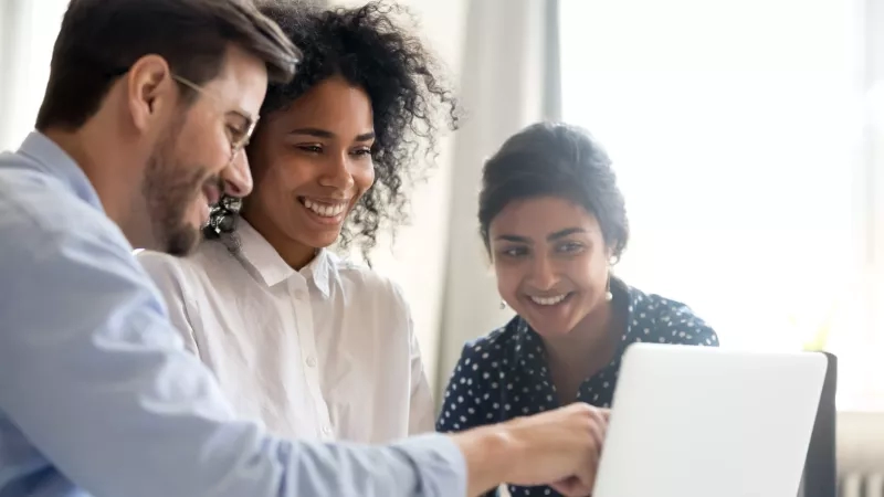Three people are sitting together with a laptop, smiling and looking at the screen of the laptop.