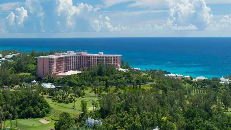 Aerial view of pink hotel by the ocean with golf course and lush trees on a sunny day.