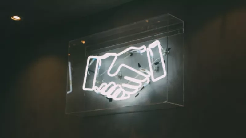Neon sign of two hands shaking in a handshake on a dark background with ceiling lights.
