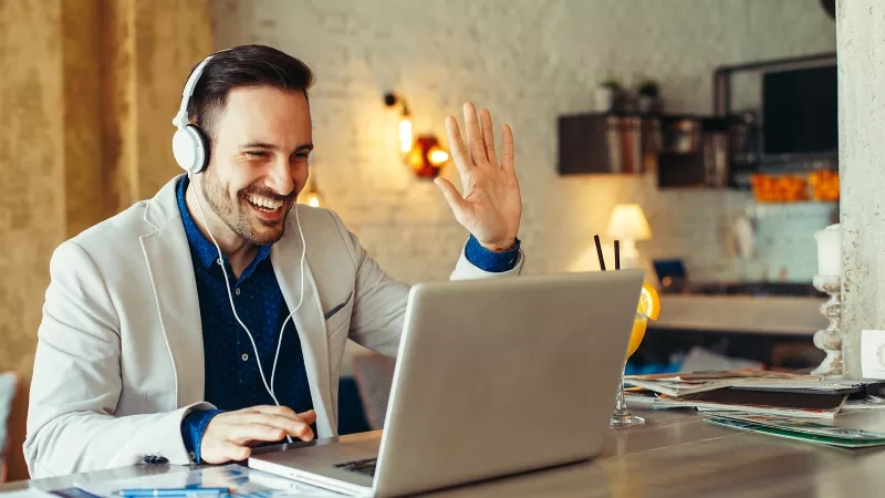 Man wearing headphones and waving hand at the camera while working on his laptop in a restaurant.