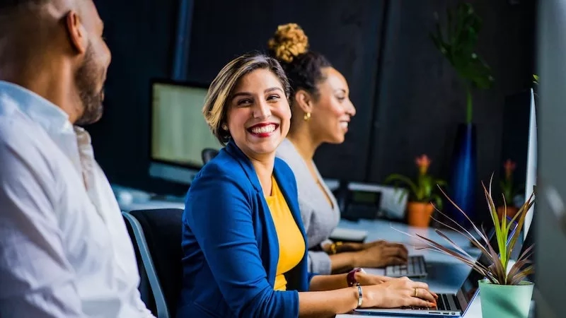 Two women and a man in an office setting, working on computers, and smiling at the camera.