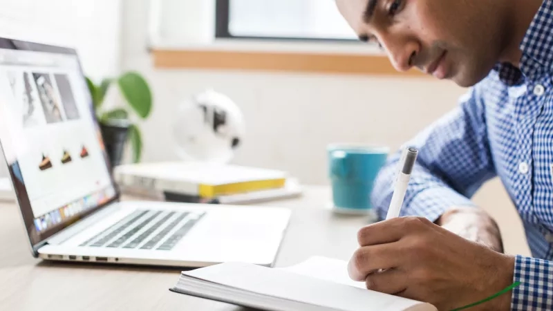 Man writing in a notebook while looking at a laptop open to an e-commerce website.