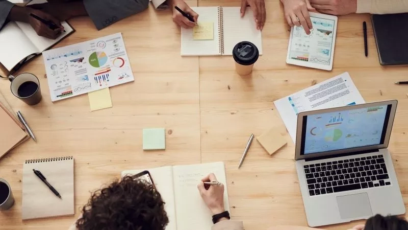 Group of people in a meeting with laptops, tablets, papers, sticky notes, and coffee cups on the table.