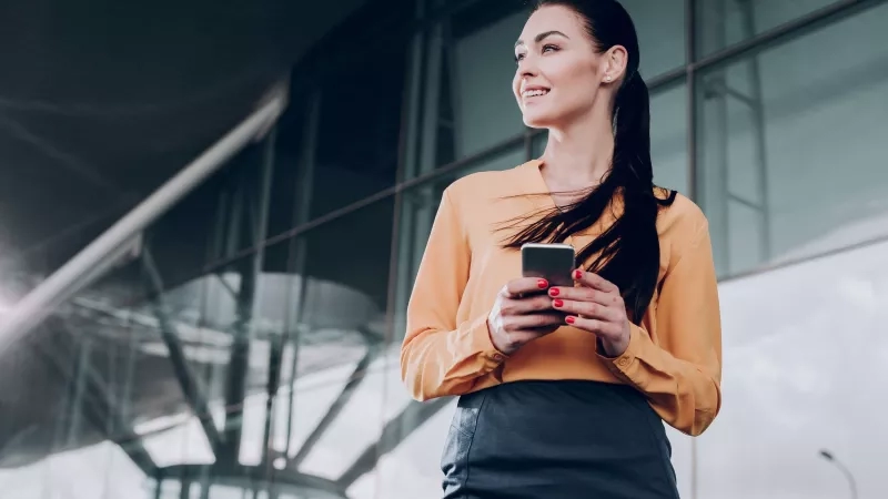 A woman smiles and looks at her phone in front of an office building.