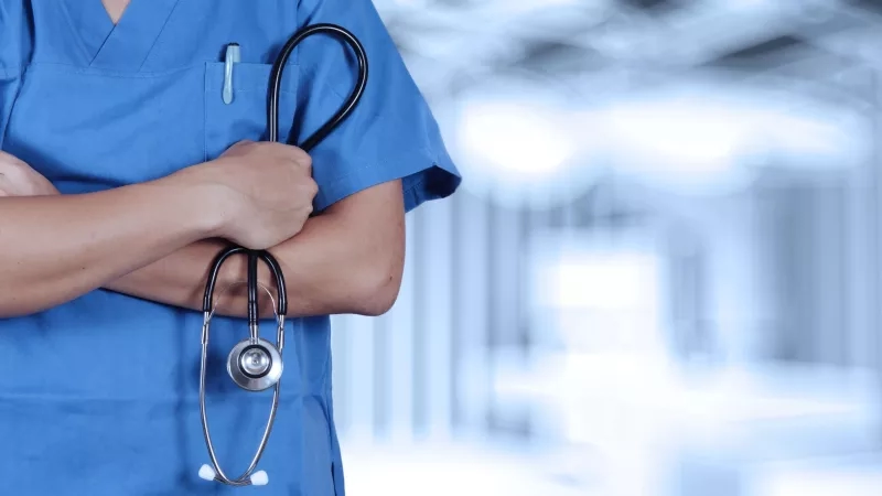 A nurse wearing blue scrubs stands with arms crossed holding a stethoscope.