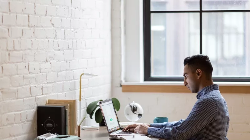 Man sitting on green chair while using laptop with white brick wall and glass window in background.