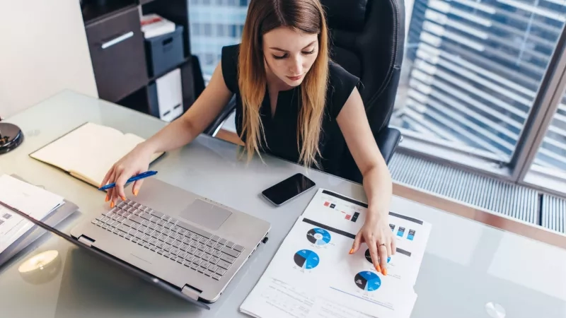 Businesswoman sitting at her office desk analyzing charts on a laptop and a paper print out.