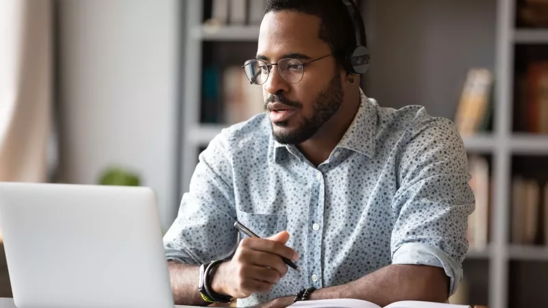 A man with headphones working from home on a laptop with a book and pen in front of him.