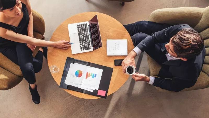 Two people sitting on chairs around a table with a laptop and documents, in an office.