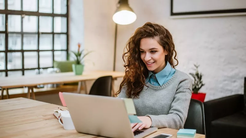 Smiling girl using a laptop in a London co-working space with sticky notes and a smartphone on a desk.