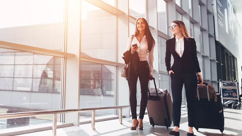 Two women are walking in the airport with their luggage and handbags.