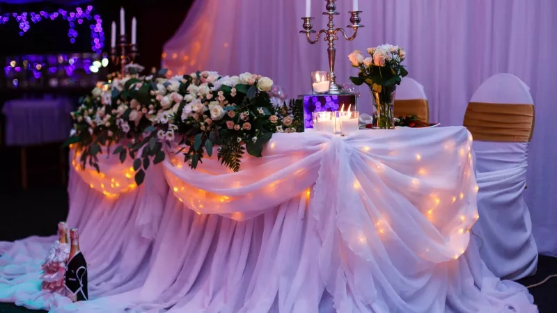 Wedding sweetheart table with white cloth, candles, flowers, and purple curtains at a banquet hall.