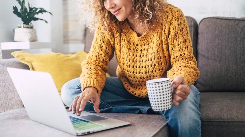 Woman sitting on a couch with a laptop, smiling while holding a coffee cup.