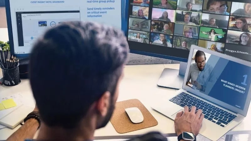 A man sitting at a desk with a laptop, mouse, and computer screen, participating in a video call.