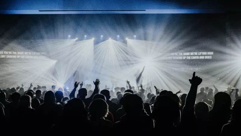 A crowd at a live event with a bright stage, with text and beams of light.