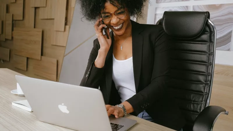 A smiling woman on the phone while working on a laptop in an office setting.