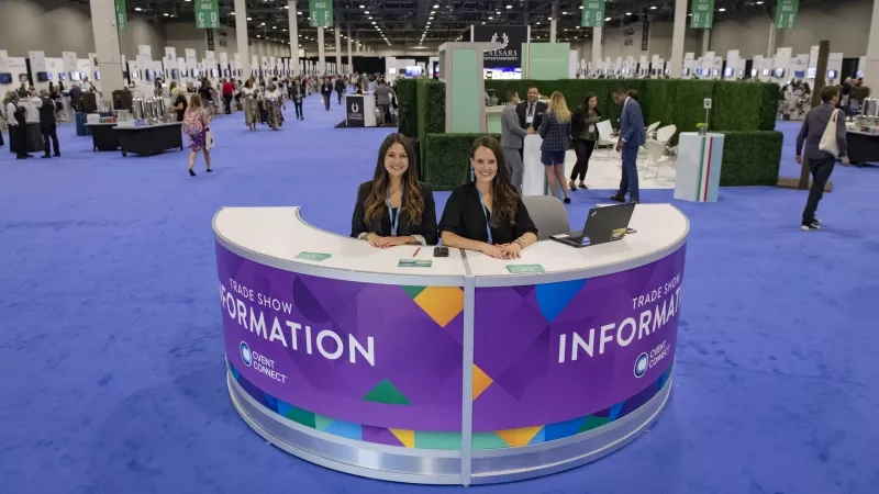 Two women sitting at a trade show information booth in a large convention center.