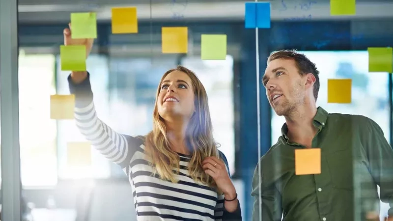 Group of marketers working together in office with post-its on glass wall and smiling at each other.