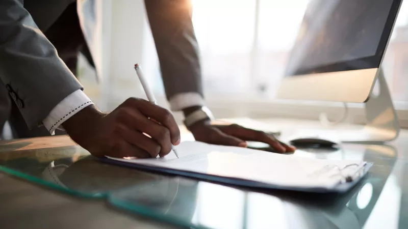 Man signing a document on a glass desk with a computer monitor in the background.