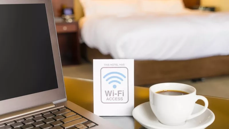 A laptop, coffee cup, and Wi-Fi access card are on a table in front of a bed.