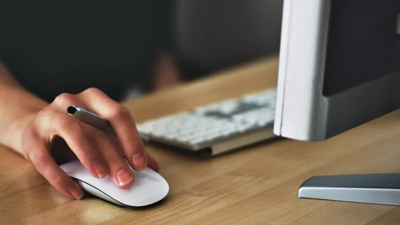 Hand using a computer mouse in front of a computer screen and keyboard in a workspace.