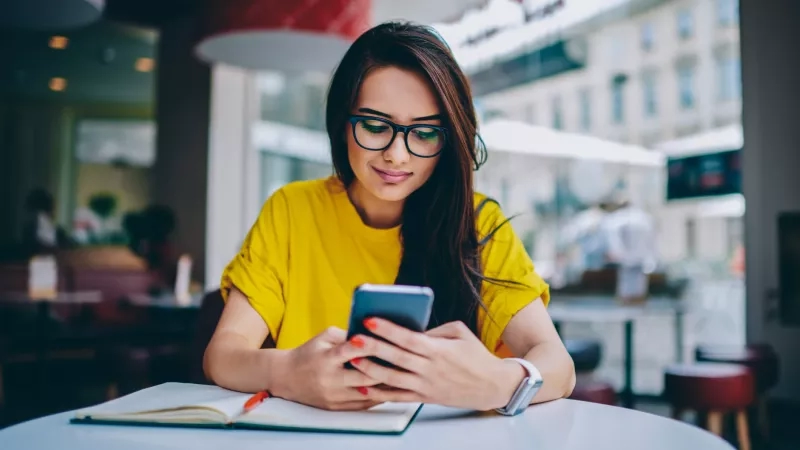 Woman sitting at a table using smartphone with book and pen in front of her.