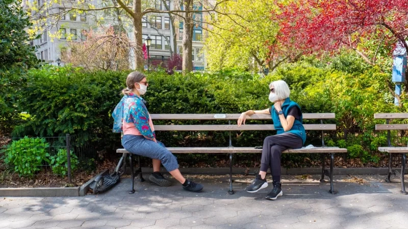 Two women wearing masks sit on a bench in a park with trees and buildings in the background.