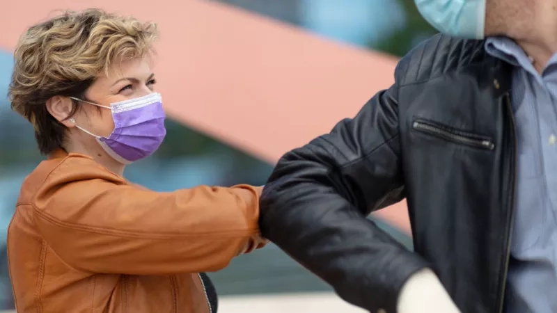 A woman wearing a mask and a man wearing gloves and a mask touch each other's arms.