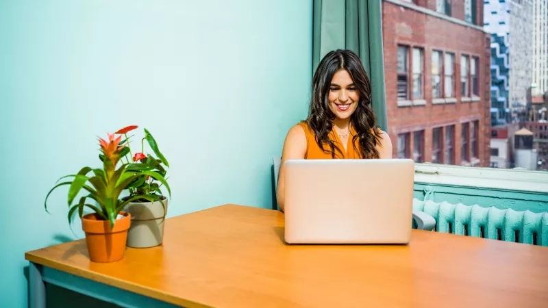 A smiling woman working on her laptop in front of a window at the Refinery.