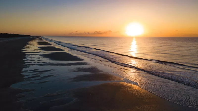 The sun sets over the horizon of the ocean, casting a warm glow on the beach.